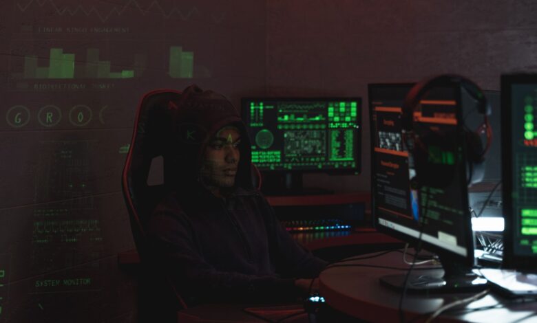 A man in a black hoodie engaged in cybersecurity work using multiple monitors indoors.