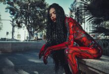 A cosplayer in a vibrant red costume sitting and posing outdoors amidst city buildings and trees.