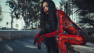 A cosplayer in a vibrant red costume sitting and posing outdoors amidst city buildings and trees.