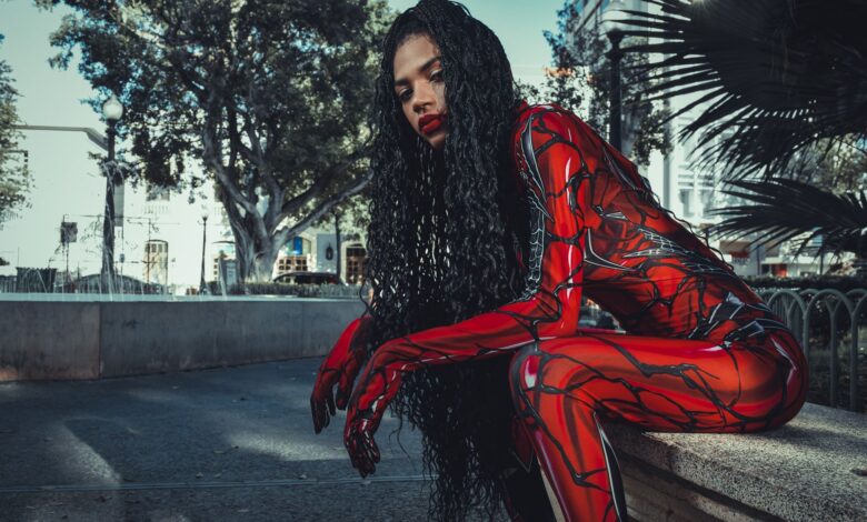 A cosplayer in a vibrant red costume sitting and posing outdoors amidst city buildings and trees.