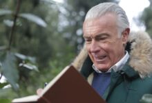 Elderly man in winter coat, writing in a notebook outdoors with a thoughtful expression.