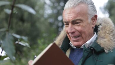 Elderly man in winter coat, writing in a notebook outdoors with a thoughtful expression.