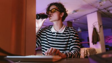 A young man with glasses drinks soda while working on his computer in a vintage office setting.