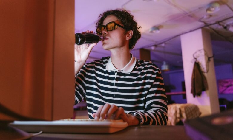 A young man with glasses drinks soda while working on his computer in a vintage office setting.