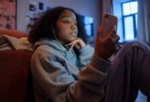 Teenage girl using smartphone indoors with earphones, wearing a hoodie, and looking relaxed.