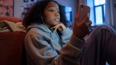 Teenage girl using smartphone indoors with earphones, wearing a hoodie, and looking relaxed.