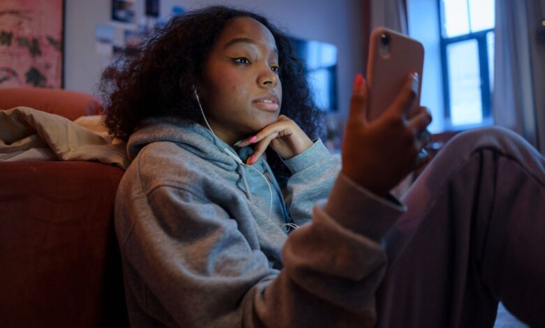 Teenage girl using smartphone indoors with earphones, wearing a hoodie, and looking relaxed.