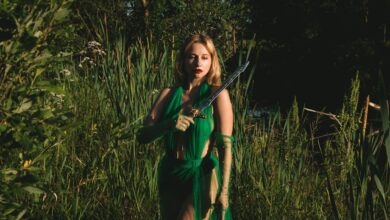 A woman in a green dress holds a sword in a mystical forest setting, evoking fantasy themes.