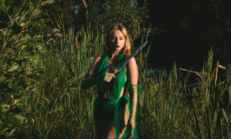 A woman in a green dress holds a sword in a mystical forest setting, evoking fantasy themes.
