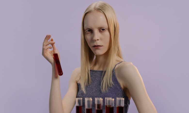 Blonde woman holding a vial with red liquid, set against a purple background.