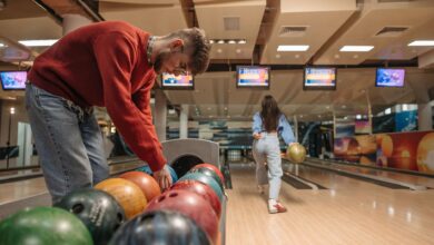 A man in a red sweater selects a bowling ball while a woman rolls a ball down the lane.
