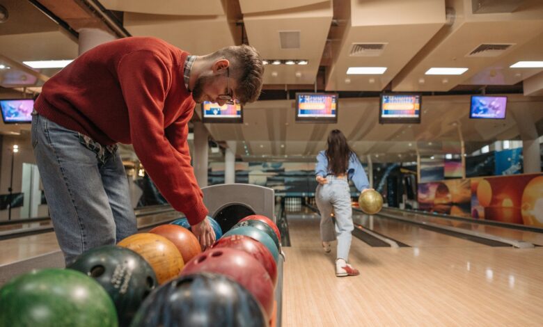 A man in a red sweater selects a bowling ball while a woman rolls a ball down the lane.