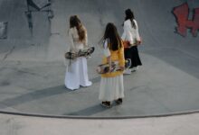 Three women skateboarders in stylish outfits strolling in a skatepark, holding skateboards.