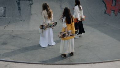 Three women skateboarders in stylish outfits strolling in a skatepark, holding skateboards.