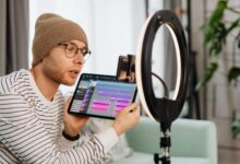 A young man wearing glasses and a beanie records a video indoors using a tablet and ring light.