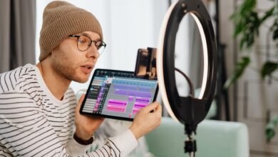 A young man wearing glasses and a beanie records a video indoors using a tablet and ring light.