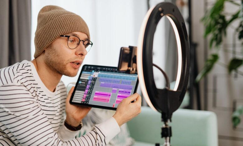 A young man wearing glasses and a beanie records a video indoors using a tablet and ring light.
