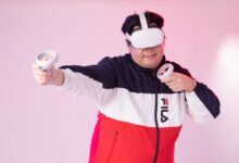 Adult man wearing VR goggles and jacket, playing with controllers against a pink studio background.