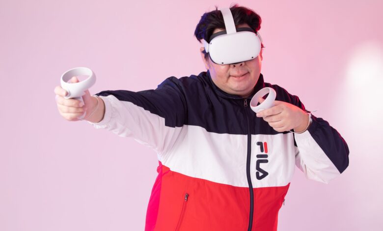 Adult man wearing VR goggles and jacket, playing with controllers against a pink studio background.