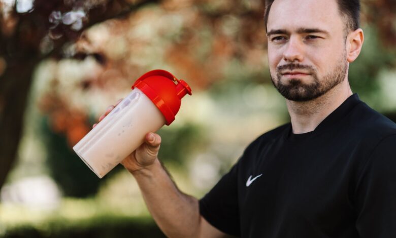 Adult man holds a protein shake in a red lid tumbler outdoors, wearing a black shirt.