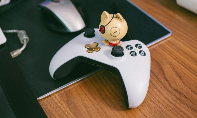 A charming cat figurine sits atop a gaming controller, beside a computer mouse, on a wooden desk.