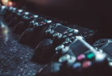 Series of gaming controllers lined up on a reflective surface, dimly lit.