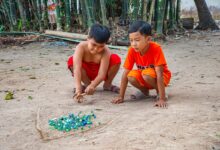 Two boys play with marbles in a sandy outdoor area surrounded by bamboo.