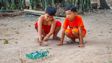 Two boys play with marbles in a sandy outdoor area surrounded by bamboo.