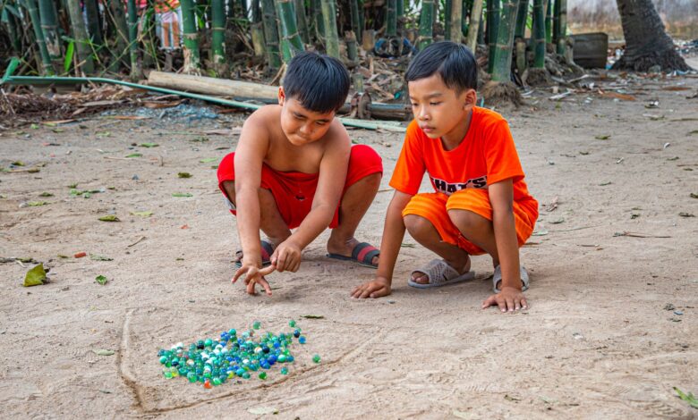 Two boys play with marbles in a sandy outdoor area surrounded by bamboo.