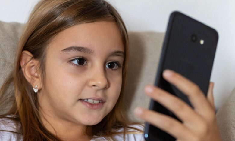 A young girl with long brown hair smiles while using a smartphone indoors.