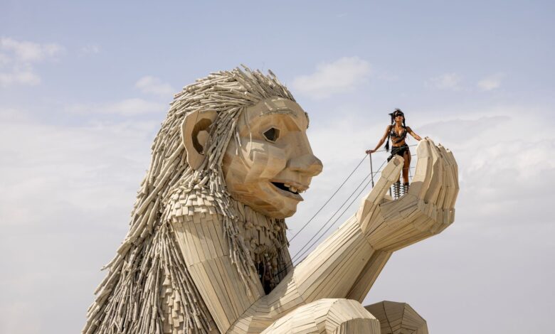 Person atop giant wooden sculpture at Burning Man festival in desert.