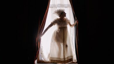 Elegant woman in Victorian attire backlit at a window with dramatic curtains.