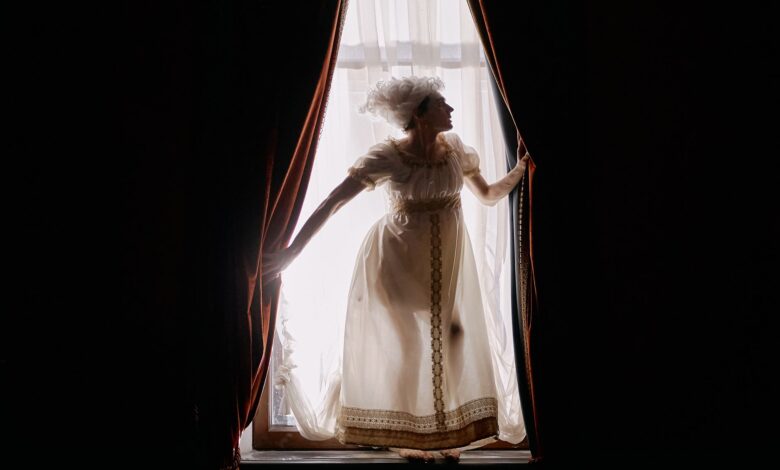 Elegant woman in Victorian attire backlit at a window with dramatic curtains.