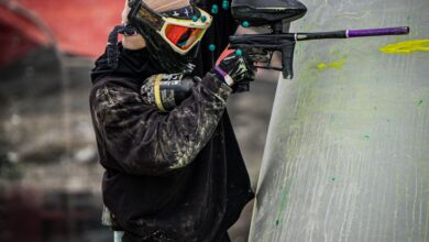 A paintball player in full gear taking aim during a competitive game outdoors.