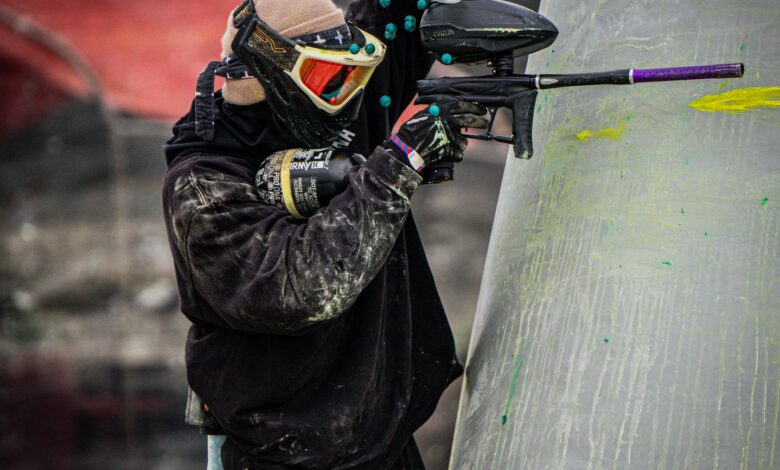 A paintball player in full gear taking aim during a competitive game outdoors.