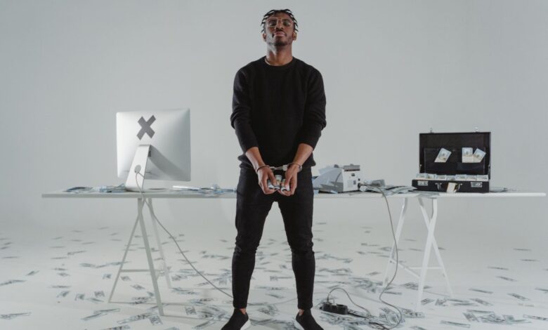 A handcuffed man stands in a room with scattered money, a computer, and a briefcase.