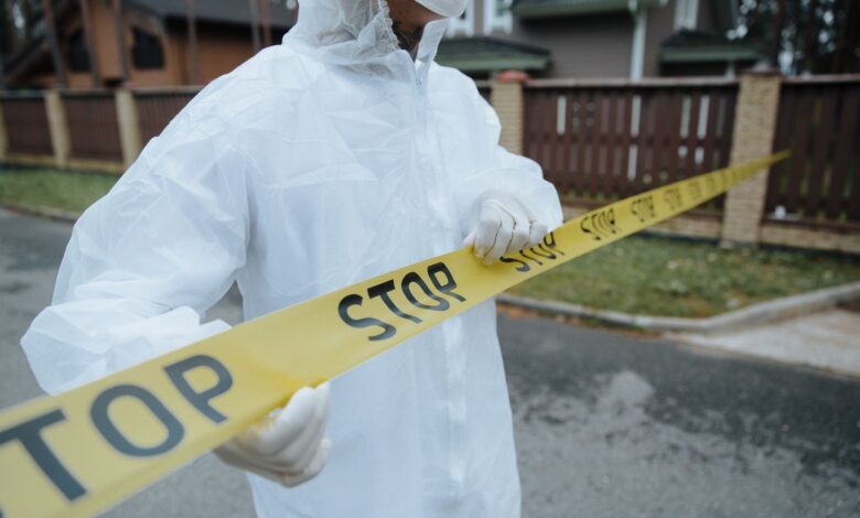 A person wearing a protective suit, handling yellow tape with 'STOP' in an outdoor urban setting.