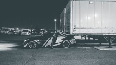 Black and white of anonymous driver getting out of modern automobile with headlight light parked on asphalt road near structure in evening