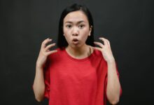 Asian woman in a red shirt expressing surprise with hands raised, on a dark background.
