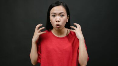 Asian woman in a red shirt expressing surprise with hands raised, on a dark background.