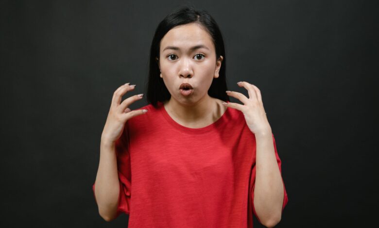 Asian woman in a red shirt expressing surprise with hands raised, on a dark background.