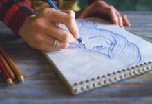 Close-up of a hand sketching a portrait on a spiral sketchpad with colored pencils on a table.