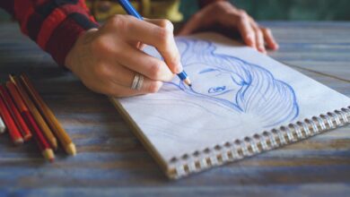 Close-up of a hand sketching a portrait on a spiral sketchpad with colored pencils on a table.