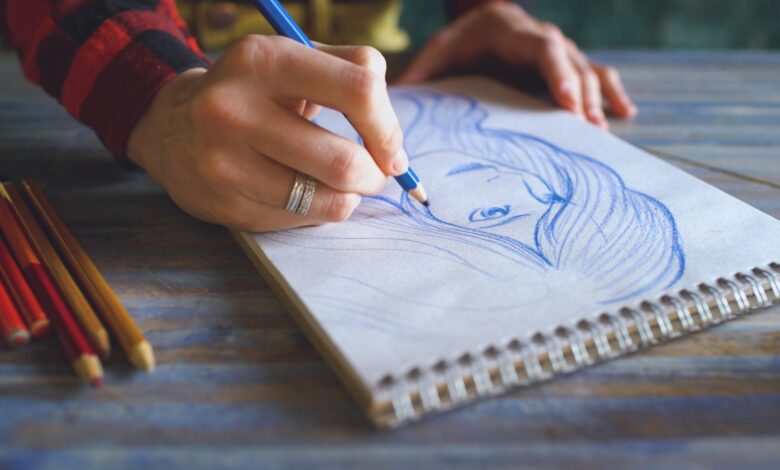 Close-up of a hand sketching a portrait on a spiral sketchpad with colored pencils on a table.