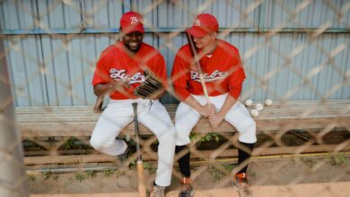 Two baseball players in red uniforms sitting on a dugout bench, chatting and smiling.