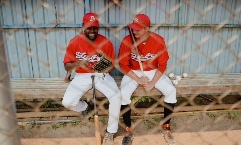 Two baseball players in red uniforms sitting on a dugout bench, chatting and smiling.