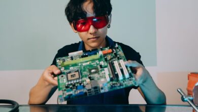 A focused young engineer holding a motherboard, wearing eye protection indoors.