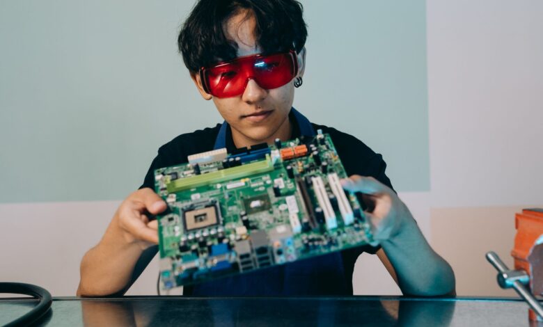 A focused young engineer holding a motherboard, wearing eye protection indoors.