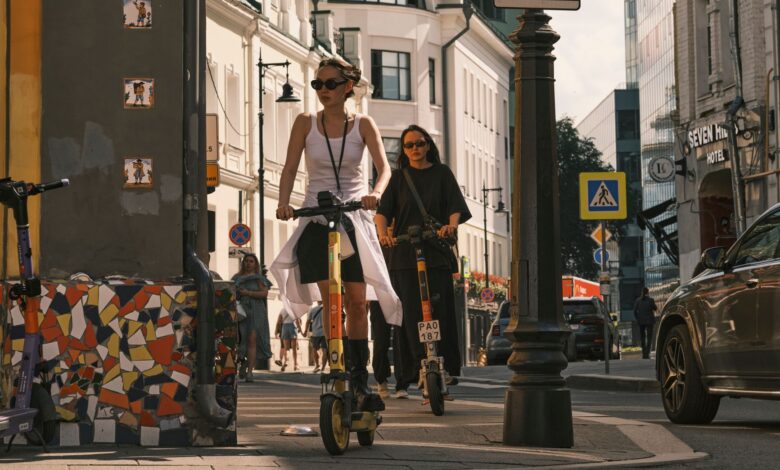 Two women riding electric scooters on a city street, showcasing urban commute.