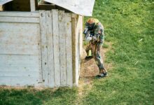 A paintball player taking cover behind a wooden structure on a grassy field during a game.